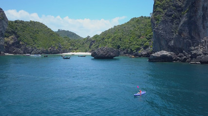 Fototapeta na wymiar AERIAL: Tiny woman in kayak is surrounded by large limestone cliffs and vast sea