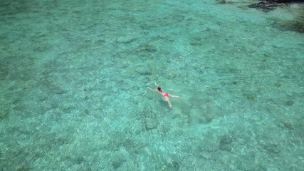 AERIAL: Flying above lonely Caucasian woman swimming in crystal clear ocean.