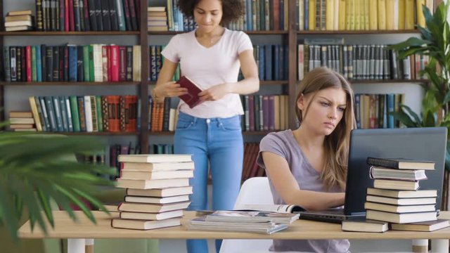 Two attractive female students caucasian blonde and african brunette take a book off the shelf in a library consult discuss and type using laptop at bureau table studying and working
