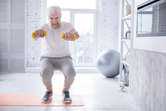 Upbeat mood. Joyful senior man doing squats and posing for the camera, smiling brightly, while holding a pair of dumbbells