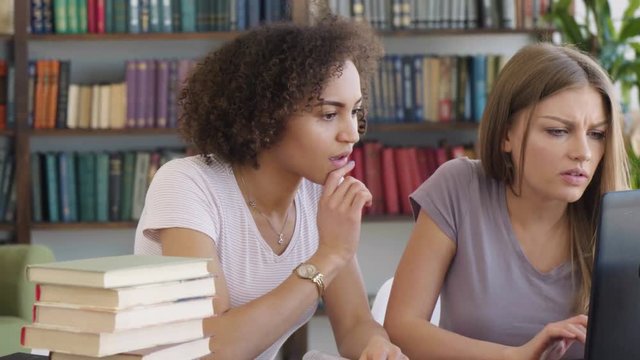 Close-up of two attractive female students caucasian blonde and african brunette consult book discuss and type using laptop studying in front of bookcase sitting at the table with books in a library