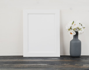 White frame, flower in the vase, clock on dark grey wooden table against the white wall with copy space. Mock up.