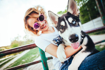 Obraz premium girl with curly hair in pink glasses sits on the bench and looks on dog with different eyes color on her hands in the spring in the park