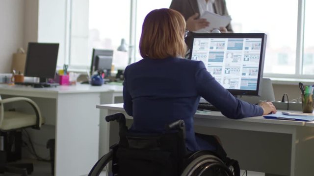 Tracking rear view of disabled businesswoman on wheelchair working on computer at office desk while her male colleague walking with documents
