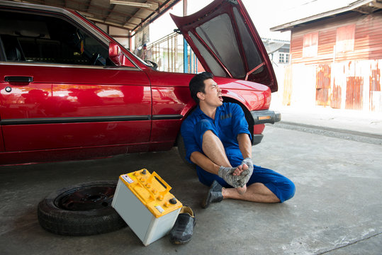 Asian mechanic get Injury and suffering at foot from battery car in garage. Foot Pain ,young man sitting on the floor at garage, he holding feet press massage. Incident case in the garage.