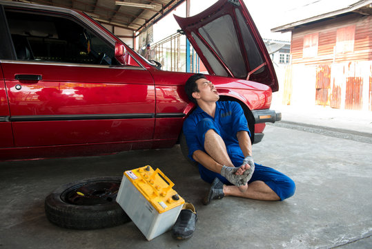 Asian mechanic get Injury and suffering at foot from battery car in garage. Foot Pain ,young man sitting on the floor at garage, he holding feet press massage. Incident case in the garage.