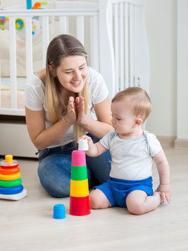 Happy young mother cheering while her toddler boy building tower from toy blocks