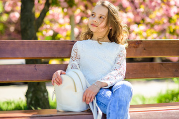 Girl on dreamy face sits on bench, sakura tree on background, defocused. Spring walks concept. Cute child with backpack enjoy sunny spring day. Girl relaxing while walk in park near cherry blossom.