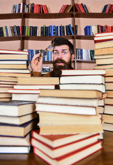 Teacher or student with beard wears eyeglasses, sits at table with books, defocused. Scientific research concept. Man on surprised face between piles of books in library, bookshelves on background.