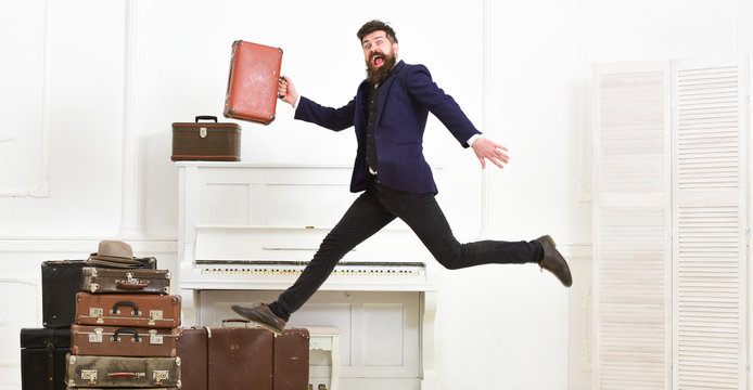 Long awaited vacation concept. Macho attractive, elegant on cheerful face carries vintage suitcases, jumping. Man with beard and mustache in suit carries luggage, luxury white interior background.