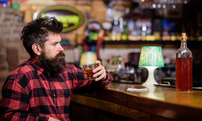 Guy spend leisure in bar, defocused background. Hipster with beard ordered full bottle of alcohol. Relaxation concept. Man drinks whiskey or cognac. Man with strict face sits near bar counter.