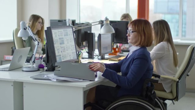 Tilt up of paraplegic businesswoman sitting in wheelchair at office desk, working with documents and typing on computer