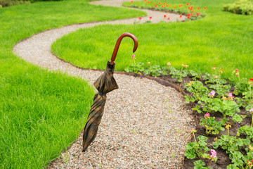 a winding footpath in the garden, a folded umbrella, green grass and flowers along the path, a concept of rainy weather