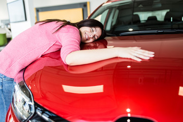 Happy beautiful young woman hugging a new car she wants to buy at the car showroom.