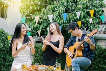 Group of friends having party in home garden.Happy young mates enjoying party playing guitar, singing and drinking beer eating food in garden.