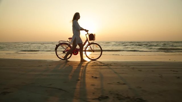 A young girl rolls her bike next to her on the beach at sunset. Silhouette of a girl with long hair. A woman in a white dress rolls a bike along the surf line.