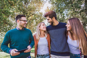 Image of four happy smiling young friends walking outdoors in the park holding digital tablet