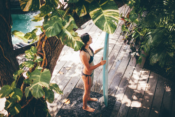 Surfer girl washing blue surfboard in the outdoor shower with tropical plants, view from above at surf camp in Bali, Indonesia