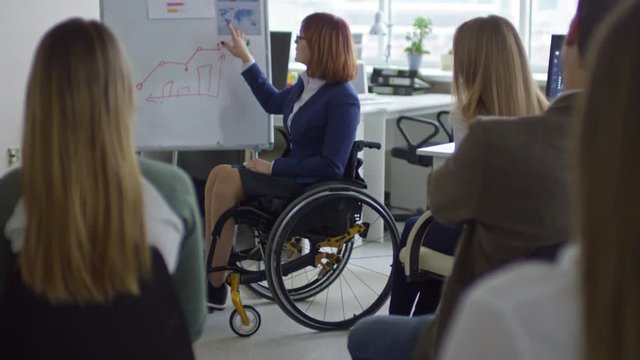 Tilt up of female business coach in wheelchair speaking to office workers and explaining bar graph on flipchart