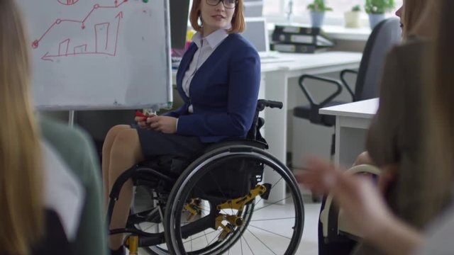 Tilt up of paraplegic businesswoman in wheelchair answering questions of group of employees and explaining graph on flipchart during business training