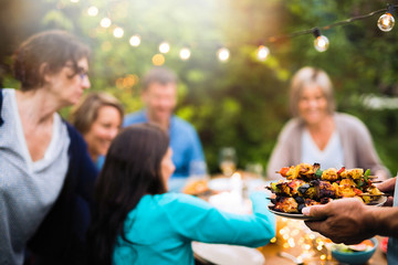  group of friends in their forties gathered around a table in the garden to share a bbq meal. A man offers chicken skewers to guests