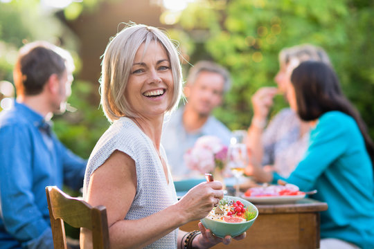 Looking at the camera a beautiful woman in her forties with a bowl of mixed salad by hand. She sits around a table in a garden with friends for dinner.