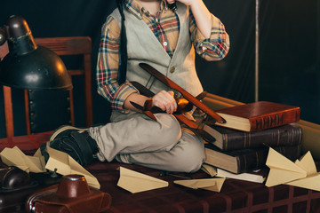 the boy is sitting on the table. near homemade airplanes made of paper, and old books, magnifier and old camera. the chair is pushed to the table.