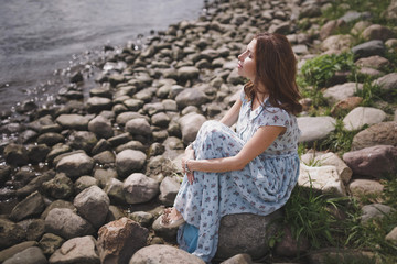 A beautiful young brunette woman with long hair in a long blue dress sits by the lake or river on a...