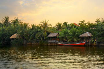 Naklejka na ściany i meble Pristine riverbank at sunset, palms trees, a red boat and bungalows, situated at the Praek Tuek Chhu River in Kampot Province in southern Cambodia, Asia. Natural asian riverside landscape