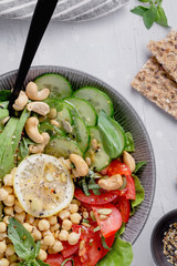 Top view of a healthy vegan lunch bowl with raw avocado, chickpeas, cashew, tomatoes, cucumber, greens and seeds over white kitchen table.