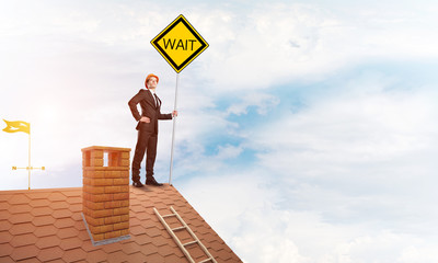 Young businessman on house brick roof holding yellow signboard. Mixed media