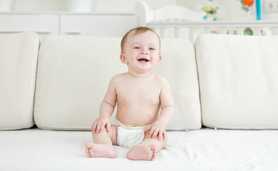 Happy laughing toddler boy in diapers sitting on sofa and looking in camera
