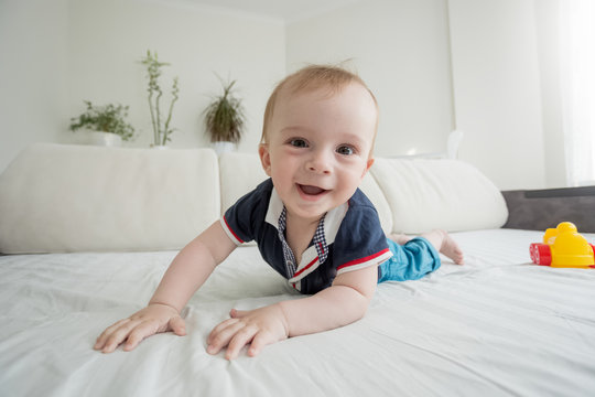 Wide angle image of happy smiling toddler boy lying on bed and looking in camera