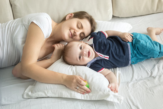 Portrait of happy smiling baby boy lying in bed next to sleeping young mother