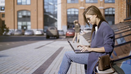 Fototapeta na wymiar A young pretty concentrated girl in jeans in sunglasses is working with a laptop outdoors near a loft