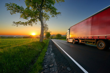 Truck driving on the asphalt road between trees in a rural landscape at sunset