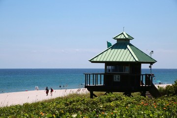 Lifeguard Station with Two People Walking on the Beach and Green Foliage Below on a Bright Sunny Day with Clear Blue Sky Overhead at Spanish River Park, Boca Raton, Florida - Powered by Adobe