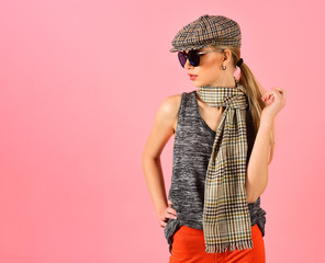 Side view skinny blond model in tartan beige cap and scarf and black sunglasses isolated on pink background. Girl in bright orange pants playing with her ponytail