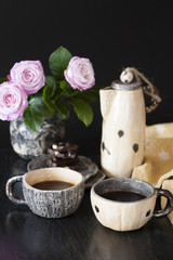 Two cups of black coffee, black chocolate, a yellow teapot and a vase with pink roses on a dark background.