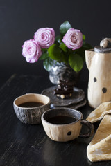 Two cups of black coffee, black chocolate, a yellow teapot and a vase with pink roses on a dark background.