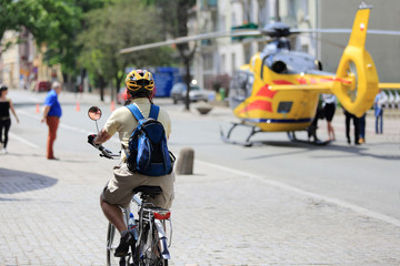 Mężczyzna jedzie rowerem po chodniku w centrum miasta obok stojącego na ulicy śigłowca pogotowia lotniczego. - obrazy, fototapety, plakaty