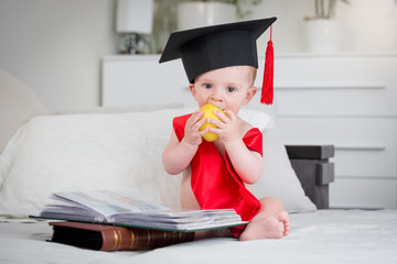 Adorable baby boy in graduation cap sitting on bed with books and biting apple