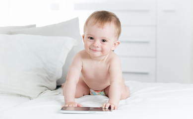 Portrait of smiling baby boy in diapers crawling towards digital tablet computer and looking in camera