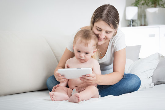 Young mother sitting on bed with her baby son and showing him cartoons on digital tablet