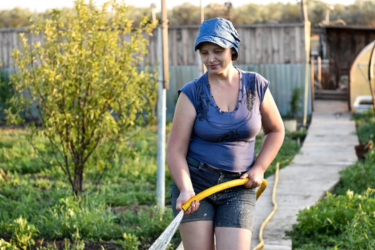 Sensual caucasian woman pours water from a hose flowers and vegetables, trees in her garden. A hot summer day, a river in the background, a greenhouse, an excellent harvest of fruits and vegetation.