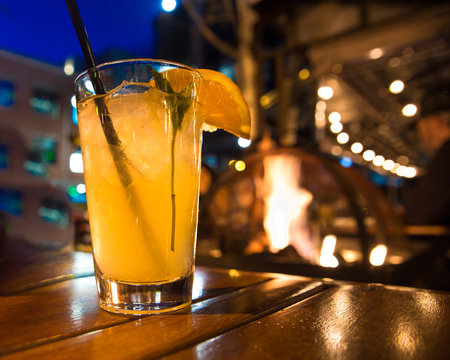 Refreshing Cocktail Beverage With Ice And Straw Scene From Outdoor Restaurant At Night With Lights And Firepit. 