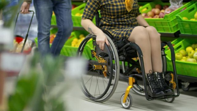 Low section of paraplegic woman riding wheelchair along produce shelves in supermarket while her husband walking behind and pulling cart with groceries