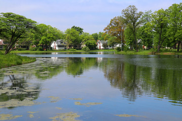 Fototapeta na wymiar Beautiful summer landscape with cloudy blue sky reflected in a lake Mendota bay water and neighborhood buildings between fresh green trees on a background. Tenney Park,city of Madison, Wisconsin,USA.