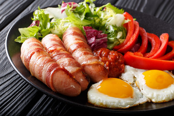Full meal: Pigs in blankets - fried sausages wrapped in bacon, eggs, sauce and salad close-up on a plate. horizontal