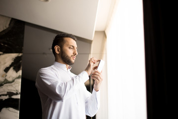 Fototapeta premium Serious guy volumizing image in smartphone while standing in hotel room by window